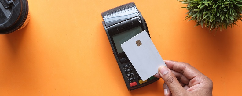 Mão segurando um cartão de crédito na frente de uma maquininha de cartão.